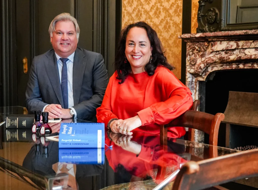 Een man en een vrouw zitten lachend aan een glimmende tafel met een wetboek