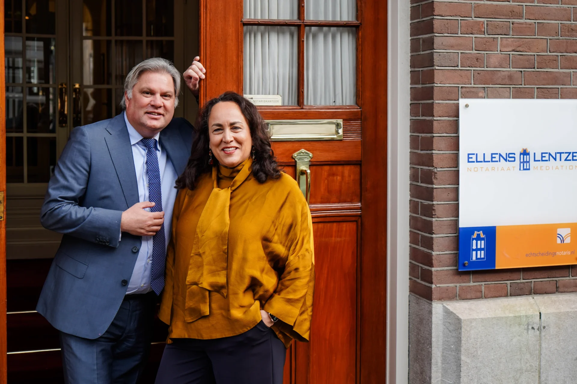 A man and a woman pose at the front door, next to the Ellens & Lentze sign