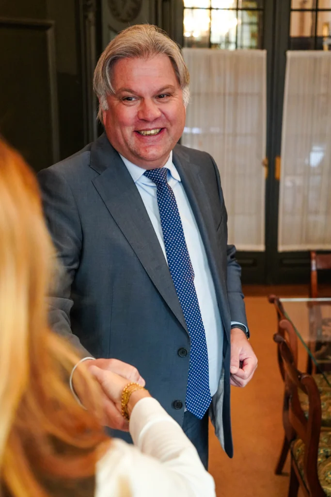 A smiling man in a suit shakes hands with a person who is half in frame