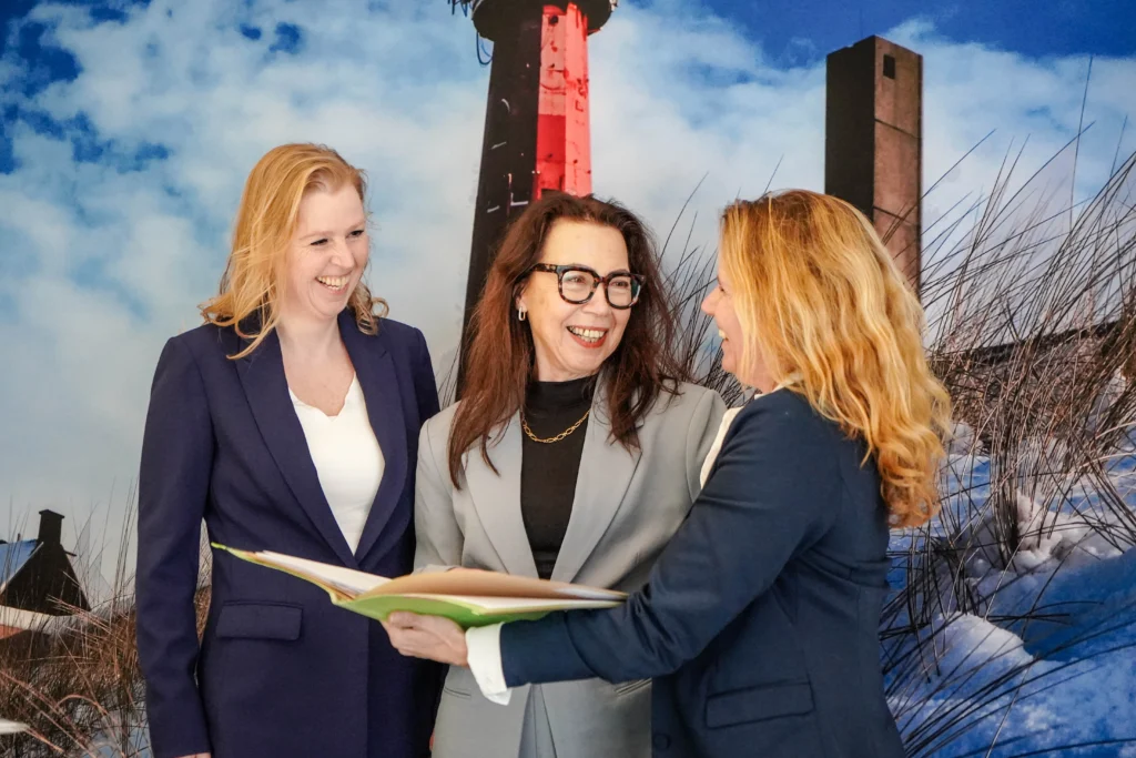 Drie vrouwen in zakelijke kleding kijken samen naar een map, met een duinlandschap en vuurtoren op de achtergrond