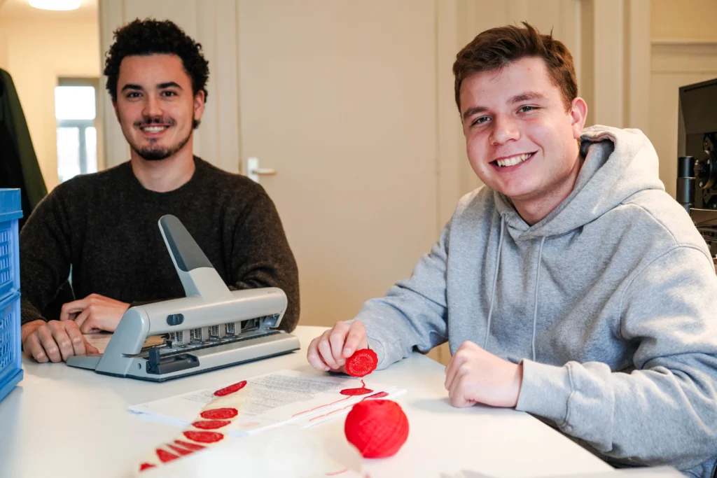 Twee jonge mannen lachen aan een tafel met een machine en rood lint met zegels